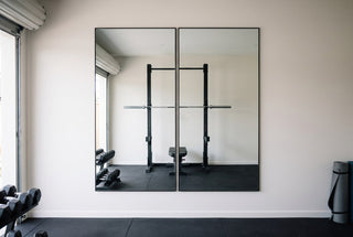 Two black framed Aldren X 200 x 100cm mirrors hung upright side by side on a white wall in a New Zealand garage gym, reflecting a squat rack, barbell and bench on black rubber flooring