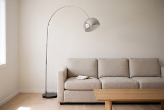 Cosmo chrome arc floor lamp with a black marble base standing beside the arm of an oatmeal linen sofa in a New Zealand lounge, its chrome dome hanging over the first seat cushion above an open book