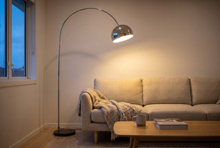 Cosmo chrome arc floor lamp with a black marble base reaching over a linen sofa in a New Zealand living room at dusk, its chrome dome glowing warm over the first seat cushion