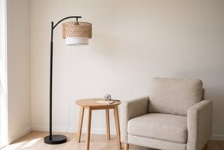 Rousseau rattan weave floor lamp standing beside a small oak side table with a tape measure and a notepad on it in a New Zealand living room, the whole lamp in frame from base to bridge arm