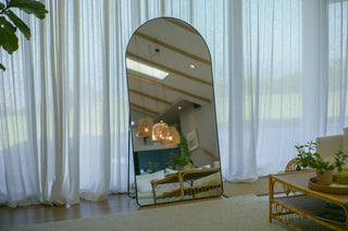 Grandeur Arched Full-Length Mirror (200×100 cm) leaning against a wall, reflecting a bright, modern living space with rattan furniture and soft curtains