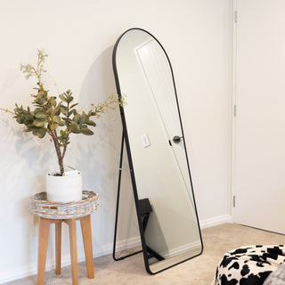 An arched full-length mirror with a black frame, leaning against a white wall in a modern, minimalist bedroom next to a small wooden stool.