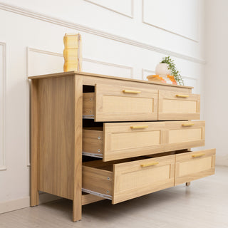 Raya 6-Drawer Lowboy dresser, 123 × 40 × 75 cm, in warm oak finish with rattan-style drawer fronts and gold handles, styled in a bright modern room with drawers open to show storage depth.