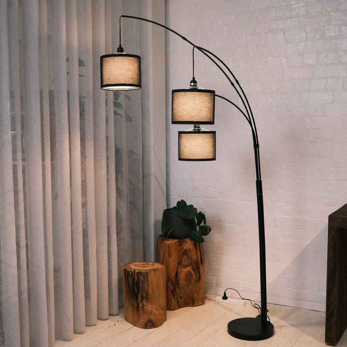Trilogy 3-light arc floor lamp featuring a matte black metal finish and curved arms. Three textured fabric drum shades are suspended at staggered heights. Shown beside rustic wooden log tables and sheer curtains. The woven shades diffuse light for a soft, warm atmosphere.