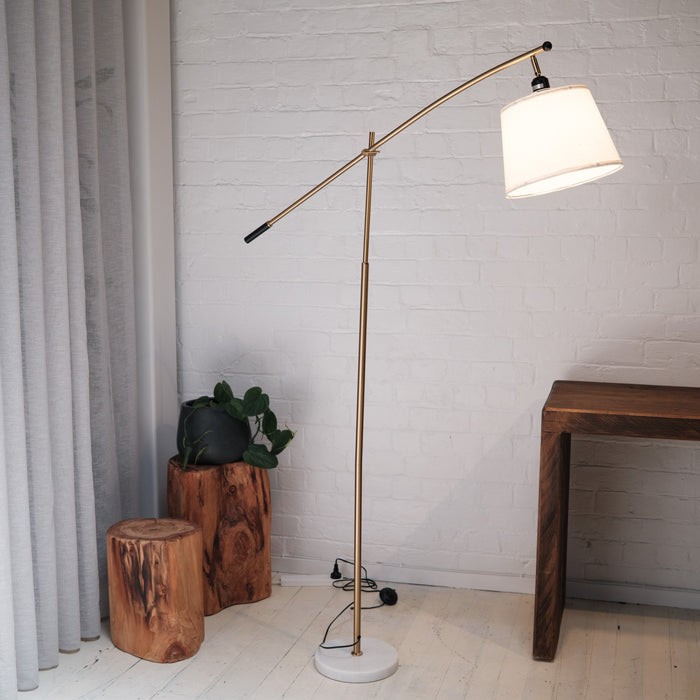 Vienna Brass Marble Base Floor Lamp styled in a contemporary living space with a white brick wall, natural timber stools, a wooden desk, and soft grey curtains. The lamp features a brass frame, fabric shade, and a solid white marble base, creating a warm and elegant atmosphere.